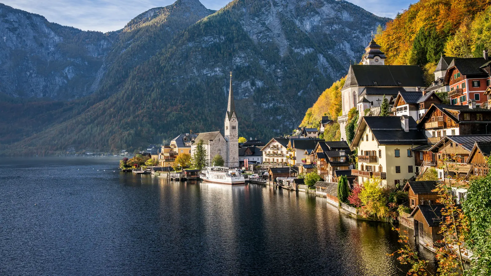 Hallstatt mit Kirchturm und Holzhäusern am Hallstätter See, dahinter ein herbstlich gefärbter Hang und Berge
